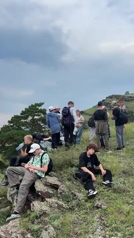 A group of young people are gathered on a grassy hillside overlooking a vast expanse of trees and a body of water. Some are sitting on rocks, others are standing and talking. One person is drinking from a bottle. The sky is cloudy, with a hint of blue peeking through. The overall impression is one of a casual outing in a natural setting.