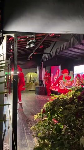 A group of people in red outfits are performing on a wooden stage. One person is wearing a colorful dragon mask. The stage is set up outdoors under a covered area with a black canopy. There are speakers and lighting equipment on the stage, as well as a large screen displaying an image of flames. Green foliage is visible in the foreground.