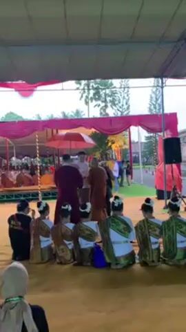 Here's a description of the scene in the video:

The scene takes place under a large tent-like structure decorated with red and white fabric. In the background, rows of individuals dressed in orange robes are seated on an elevated platform. Several other people are standing and milling about nearby. 

In the foreground, a group of young girls dressed in traditional costumes are kneeling on what appears to be a stage covered with a yellowish fabric. Their costumes consist of white tops with green and gold embellishments and their hair is decorated with white flowers. They are facing the back.

Two monks, one wearing a maroon robe and the other a lighter orange robe, are walking across the stage. A man in a white shirt is walking alongside them, holding a red umbrella over the maroon-robed monk. To the right, another individual wearing black clothing and a yellow lanyard is holding what appears to be a musical instrument.

The entire scene suggests a religious or cultural ceremony taking place outdoors. In the bottom left of the frame, a person wearing a hijab is partially visible, capturing the event with a camera.