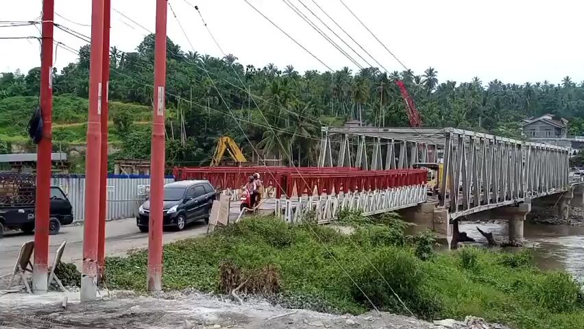 A motor vehicle is seen traveling across a bridge that is under construction. The bridge appears to be a suspension bridge with metal trusses. The road is paved and appears to be a two-lane road. There are several vehicles on the road, including a truck, a car, and a motorcycle. There is also a construction crane in the background. The environment is tropical, with palm trees and lush vegetation visible. The weather is overcast.