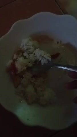 The video shows a close-up, overhead shot of a white, scalloped-edge bowl filled with rice and what appears to be some other food items. A silver spoon is visible, resting in the bowl and seemingly in the process of scooping up some of the rice. The lighting is dim, with the bowl and its contents illuminated by a soft, yellow light. The background is dark, with a hint of brown, possibly a wooden surface or tile. The overall impression is of someone eating a meal.