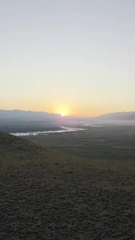 The video shows a time-lapse of a sunrise over a valley. In the foreground, there is a grassy hillside. In the middle ground, a river winds through a green valley. In the background, mountains are visible, shrouded in mist. The sun is rising on the horizon, casting a warm glow over the landscape. The sky is a pale blue, transitioning to yellow and orange near the sun. The overall atmosphere is peaceful and serene.