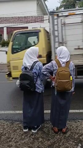 Two young women wearing white hijabs, blue patterned blouses, and gray skirts stand at a curb waiting to cross a street. One wears a black backpack and the other wears a brown backpack. They have their arms linked. A white building with red trim is visible across the street. Cars and motorcycles pass by. The weather appears cloudy and overcast.