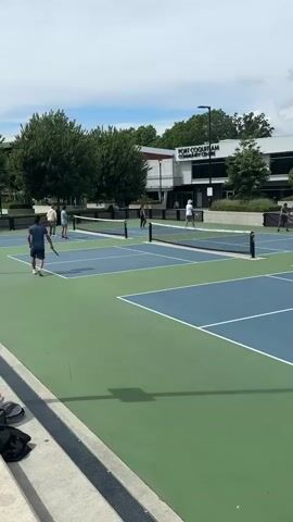 The video shows several people playing pickleball on an outdoor court with green and blue surfaces. Each court is divided by a net. There are players on two of the courts visible in the video. The backdrop includes a building with "Port Coquitlam Community Centre" written on it. Trees and a partially cloudy sky complete the outdoor scene. Players are actively hitting the ball and moving around on the courts.