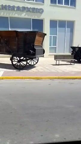 The video captures a street scene. In the foreground, a grey asphalt road is visible, with a yellow curb separating it from a paved sidewalk. A dark-colored steamroller-like object is parked on the sidewalk. Behind the steamroller, a modern white building with large windows stands. A bench is placed on the sidewalk in front of the building. Further down the street, a traffic light with red and green signals is visible. Several cars are parked on the side of the road and a white car with yellow and red markings, possibly a taxi or police vehicle, is driving away from the camera. The sky is blue with scattered clouds. The scene appears to be in an urban or semi-urban setting on a sunny day.