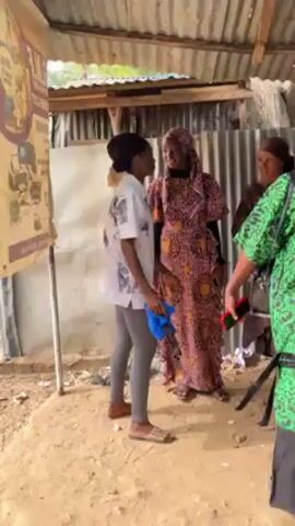 Three women are gathered outdoors in a shaded, semi-enclosed area under a corrugated metal roof. The environment appears to be simple, with sandy ground visible. 

The woman nearest the camera wears a green patterned top with a black bottom, and a small black purse. She is gesturing and speaking. Another woman arrives wearing a light blue dress with a matching headwrap and is holding a dark blue garment. The first woman briefly interacts with her.

The second woman wears a white patterned shirt with grey leggings and flats. A third woman is wearing a pink and brown patterned dress and a headscarf, standing behind the second woman. There is a poster attached to a wooden frame on the left side of the scene.