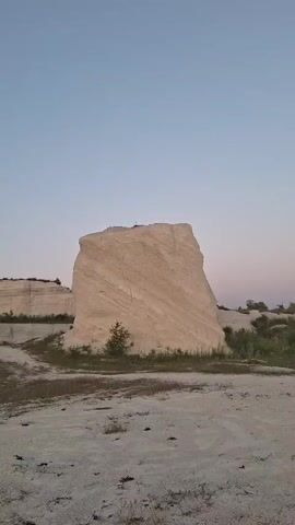 The video begins with a static shot of a large, light-colored rock formation with layered textures against a clear, pale blue sky.  In the foreground, there is dry, sandy ground with sparse, dry grass. The camera then pans to the left, revealing another section of similar rock formations.  A young woman with long, dark hair is walking away from the camera, wearing a short, dark skirt and a white top, with a patterned scarf or blanket draped over her arms. She is barefoot. The camera then moves to follow her as she walks towards a prominent, taller rock formation that has a single tree growing on its summit. The woman stops in front of this formation, holding the colorful scarf out to her sides, appearing to pose for a photo. The sky remains clear and pale.
