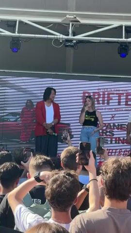The video shows a panel or presentation taking place outdoors, likely during the day given the bright lighting. Two individuals, a man and a woman, are standing on a stage in front of a large screen displaying promotional material for a film. The man, wearing a white t-shirt and a red jacket, stands to the left, holding a microphone in his right hand. The woman, in a black tank top and blue jeans, stands to his right, also holding a microphone and speaking.

The screen behind them prominently features the word "DRIFTER" in large pink letters, suggesting it's the title of the film. Other text on the screen is partially visible, including "UN FILM DI SUNG KANG" (A film by Sung Kang) and "UNA NUOVA OCCASIONE" (A new occasion). There are also images of a red car and a person in a striped outfit on the screen.

A crowd of people is gathered in front of the stage, many holding up their mobile phones to take pictures or record the event. The atmosphere appears lively and engaged. The presence of a stage, screen, microphones, and an audience strongly indicates a promotional event, perhaps a Q&A session or a premiere announcement. The location context of Cesena, Italy, suggests this event is taking place within that city.