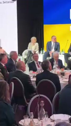 The video shows a panel discussion taking place on a stage in front of a seated audience. The backdrop is a screen displaying the Ukrainian flag. On the stage, several individuals are seated on chairs. The central figure is a woman with short blonde hair, wearing a light-colored blazer and dark pants. She is looking towards the audience. To her right is a man in a blue suit and glasses, also observing the audience.  

Below the stage, the audience is seated at round tables set with plates, silverware, and bottles of water. The majority of attendees are wearing dark suits, and they are focused on the panel discussion. A few individuals in the foreground are partially visible, including a person with glasses in a green and camouflage jacket.

The overall setting appears to be a formal event or conference, likely related to discussions about Ukraine.