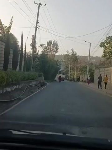 A car drives down a paved road. On the right sidewalk, a man walks ahead, and a woman in pink walks in the opposite direction. In the distance, a motorcycle follows a red car up the road toward a building. Power lines crisscross overhead amidst trees. The sky is overcast.