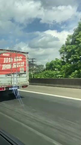 A video recorded from a moving vehicle shows a red and black box truck with Chinese text on its side driving on a highway. The truck appears to be a delivery vehicle. The video also captures trees, a cloudy sky, and a concrete barrier along the side of the road. The camera perspective is from inside the vehicle looking out the side window.