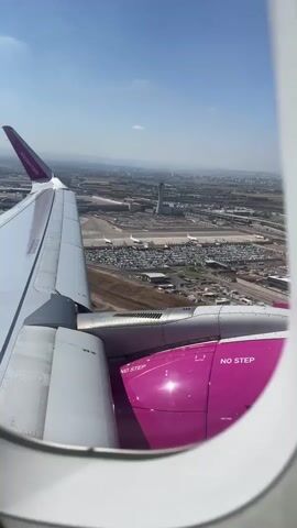 The video, taken from an airplane window, shows an aerial view of an airport and its surroundings on a clear, sunny day. The left side of the frame is dominated by the white wing of the airplane, with the purple tip and a logo visible. Below the wing, the engine housing, painted a vibrant magenta, is clearly visible, with the text "NO STEP" printed in white on its surface.

The view outside the window reveals a sprawling airport complex. A large parking lot filled with cars is a prominent feature, situated near airport terminals and other airport infrastructure. A control tower stands tall in the distance, and railway tracks run through the landscape. Beyond the airport, a cityscape stretches out, with buildings and roads indicating a populated area. The terrain appears relatively flat, with some hills or mountains visible on the horizon. The sky is a clear blue with a few wispy clouds. The airplane appears to be either taking off or landing, given the proximity to the ground and the view of the airport.