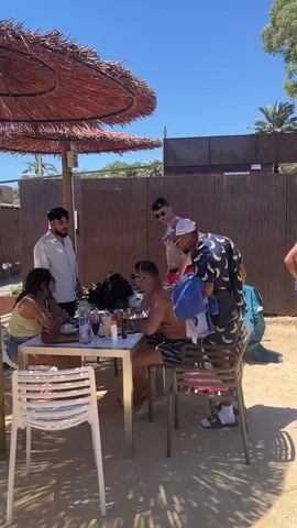 The scene is outdoors on a sunny day with a group of people gathered near a sandy area. A table with food and drinks is set up under a straw umbrella. Several individuals are seated at the table, while others are standing nearby. One person appears to be taking photos with a camera. Near a corrugated metal fence, there's a paddleboard, tires, and various bags. People are interacting and relaxing. The overall atmosphere seems casual and beach-like.