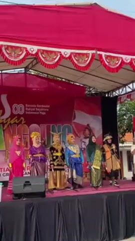 A wide shot shows seven people on a stage under a red canopy decorated with pennants.  A banner at the back of the stage is red and white, with an Indonesian flag visible. The people are women in colorful, traditional-style costumes. Each woman holds a microphone. They appear to be singing. Two people hold Indonesian flags. One stands stage right and the other left of the women. A speaker is at the front of the stage, to the left. The sky is bright blue, and the environment is outdoors.