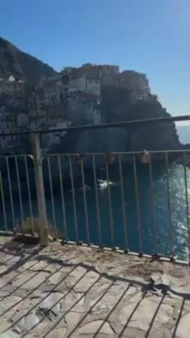The video shows an outdoor scene with a view of a coastal town built on a steep hillside. The town is characterized by closely packed, colorful buildings. In the foreground, there is a stone-paved walkway with a metal railing overlooking the sea. Several people are present on the walkway. A woman with long, dark hair, wearing a patterned white and black blouse and black jeans, is standing near the railing. Another woman in a plaid coat is looking at her phone. A man in a red shirt and black jacket is walking in the background, carrying a white bag. Another person is using a phone on a tripod to film the scene. The day is sunny, and the sky is clear. The water is deep blue.
