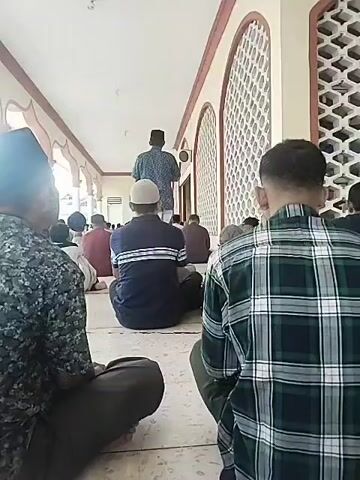The video shows a group of men sitting on the floor of a hallway in what appears to be a mosque. They are arranged in rows facing forward, listening to a man standing at the front. The speaker is wearing a patterned blue shirt and a black cap. The men are wearing a variety of casual clothing. The hallway has a series of arched windows on one side and columns on the other. The floor is tiled and the lighting is bright. The camera appears to be held by one of the men in the audience, as they are included in the frame and slightly out of focus. The overall impression is that they are participating in a religious activity or gathering.