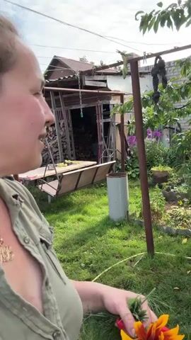 A woman is in an outdoor setting, possibly a backyard, as indicated by the presence of a swing set, a shed filled with firewood, and various plants. The woman is holding a bunch of green herbs in one hand and a bright orange and yellow flower in the other. She is wearing a green collared shirt and a gold necklace. The background shows a rustic shed with a corrugated roof, clothes hanging on a line, and a bicycle leaning against a structure. The lighting suggests it is daytime. The woman is looking towards the camera and appears to be speaking or interacting with the viewer.