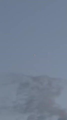 A lone aircraft flies high in a hazy, overcast sky. The aircraft is small in the frame, appearing as a speck against the pale blue-gray backdrop. Below the aircraft, wispy clouds or fog obscure the ground, suggesting a layer of atmospheric obscurity. The lighting is diffuse, indicating either early morning, late evening, or an overcast day. There is no visible text in the frame. The scene evokes a sense of isolation and the vastness of the sky.