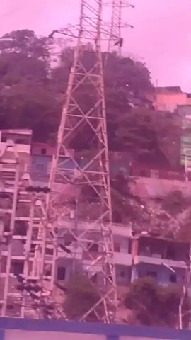 The video displays a collection of high-voltage electricity pylons situated on a hillside. The pylons are large, latticed metal structures supporting power lines. Below them, a dense collection of buildings, likely houses, is visible, clustered together on the uneven terrain. The overall color palette of the video is dominated by pink and purple hues, possibly due to lighting conditions or image processing. The scene suggests an urban or semi-urban environment where infrastructure for electricity distribution is integrated with residential areas.