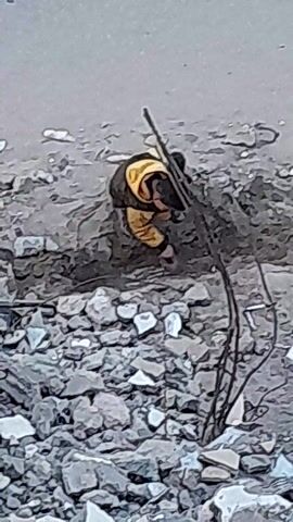 A person is visible in an overhead shot, wearing a black hat, dark pants, and a yellow jacket. They are situated amidst rubble and twisted metal debris, possibly engaging in some activity within the disrupted environment. The background comprises a mixture of gray and white tones, suggesting a demolition or damaged area.