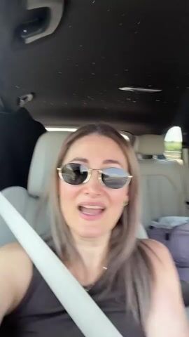 A woman wearing sunglasses and a dark tank top is sitting in the passenger seat of a car. The car's interior is visible, including the headliner which appears to have tiny lights. A white seatbelt is across her chest. Her mouth is open and her eyes are wide as she speaks to the camera. The car appears to be in motion, indicated by the slight blur of the background outside the car windows. Luggage is visible in the back seat.