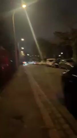 The video shows a street at night. Cars are parked along the street. Streetlights illuminate the scene. A tall building is visible in the background, and trees line the street. The camera moves along the sidewalk.