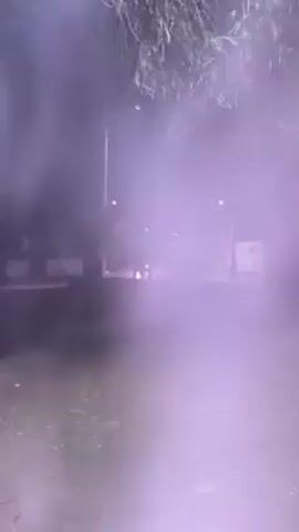 The video depicts a scene at night.  At the far end of a street, a vehicle emits bright, multi-colored lights (red, yellow, green). The street has streetlights, trees, and low buildings, and there is some sort of haze or cloud of colored smoke obscuring parts of the view. The camera appears to be handheld. The ground near the camera has dirt and leaves.