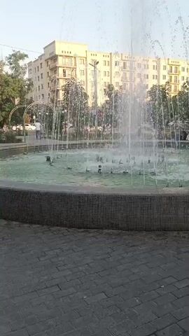 The video shows a circular fountain with multiple jets of water spraying upwards. The fountain is surrounded by a dark tiled border, and beyond that, a paved walkway with a brick pattern. In the background, there are trees and a multi-story building with many windows. A few people are visible walking in the distance, and there are also some decorative arches. The lighting suggests it is daytime.