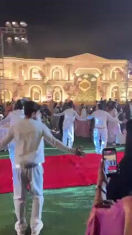 The video shows a group of men dancing at an outdoor event, possibly a wedding. They are dressed in white outfits with silver belts and white sneakers. They are dancing on a red carpet placed on a grass lawn. Behind them, there is a large, brightly lit building that appears to be part of the event venue. A crowd of people are watching, and some are recording with their phones. The event is taking place at night, and there are spotlights illuminating the scene.