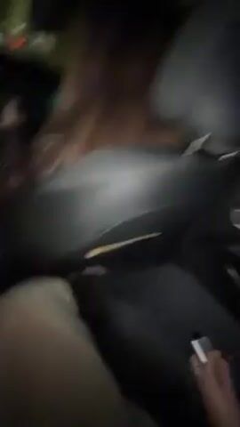 The video shows three women inside a car, likely at night. The first woman, with light skin and light brown hair, is holding a large red plastic cup, possibly a drink with a red rim. The camera shifts to show two other women, one with dark hair and another with curly brown hair. They are smiling and laughing, interacting closely. The woman with curly hair is wearing a black leather jacket. At one point, the woman holding the cup spills some of its contents, potentially onto herself or the car interior. The car appears to be moving as indicated by the background lights visible through the window.