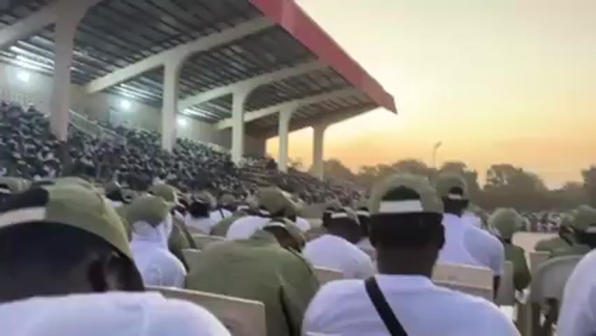 A stadium filled with people, wearing what appears to be similar green uniforms, are seated in rows of white chairs. The seating is spread out over a large field. Many people are also sitting in the bleachers, underneath a partially covered roof of a stadium. The sky overhead is shades of pink, orange, and yellow, indicating it is either dawn or dusk.
