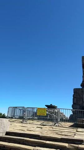 The video captures a view of a stone monument, possibly a memorial or historical landmark. The monument is large and constructed from rough-hewn stones. A weathered stone staircase leads up to a platform at the base of the monument. A metal railing is visible in the foreground, and a yellow caution sign is attached to it. The monument features a large stone sculpture of a lion in a reclining pose. Further in the distance, a scenic view of rolling hills and trees under a clear blue sky is visible. Sunlight shines brightly, casting shadows. There are no people visible in the video.