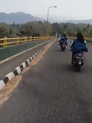 A first-person perspective video shows a group of people riding motorcycles on a road that appears to be a bridge. The road is asphalt and has a yellow and black striped curb along the right side. There are a few motorcycles visible in front of the camera, with riders wearing dark clothing. The motorcycles are moving forward. On the left side of the road, there is a yellow guardrail. In the background, there are trees, hills, and a cloudy sky. The overall environment appears to be rural or semi-rural.