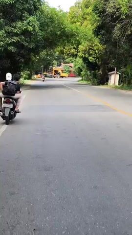 Two people ride a scooter down a paved road lined with lush green trees. The person in front is wearing a white helmet and a backpack. The person behind is wearing a dark shirt and light-colored pants. In the distance, construction equipment, including what appears to be a yellow excavator, is visible. The road has a yellow dividing line. The sky is overcast.