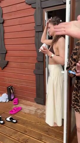 The video shows women on the porch of a red wood cabin. In the first scene, a woman with curly brown hair, wearing a brown bikini top and cream-colored skirt, stands in the doorway using a hairbrush and holding a white device. A young girl wearing a red life jacket walks by her. The next scene shows a woman in a pink robe holding a phone, and another woman holds a bottle of champagne and they both smile. The cabin is numbered 13. There is also a shopping bag, clothes and sandals on the wooden porch. The weather is overcast.