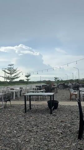 The video shows an outdoor seating area with many tables and chairs. The ground is covered in gravel. String lights are hung above the tables. In the background, there are some trees and a fence. Several people are visible, some walking and some sitting at tables. The sky is cloudy.
