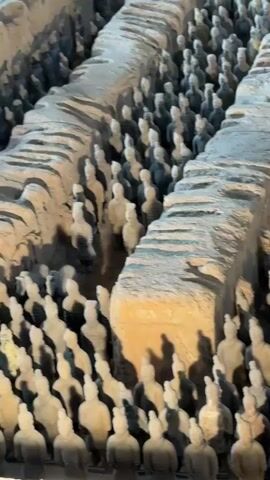This is a close-up, high-angle, panning shot of a vast army of terracotta warrior figures. They are arranged in multiple rows and columns, standing in trenches or pits. The figures are weathered and appear to be made of clay, with some retaining faint traces of color. The lighting casts shadows, highlighting the depth and scale of the formation. The overall impression is one of an ancient, meticulously crafted, and immense military force.