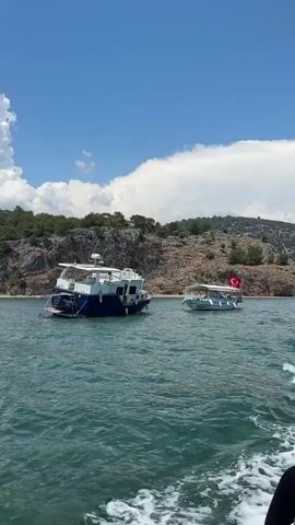 This media file depicts a scenic view from a boat on a sunny day. The water is a vibrant turquoise, and the sky is a clear blue with fluffy white clouds. In the background, a rocky, tree-covered coastline stretches out, suggesting a natural and possibly remote location.

Two boats are visible in the water. The larger boat, in the foreground, is blue and white with a cabin structure. It appears to be anchored. A smaller, lighter-colored boat is positioned further away, and it displays a Turkish flag, indicating the geographical context of Köyceğiz, Turkey. The presence of these boats suggests recreational activity, possibly a tour or a private excursion.

The overall atmosphere is serene and peaceful, with the gentle movement of the water and the vastness of the sky contributing to a sense of tranquility. The time of day appears to be midday or early afternoon, given the bright sunlight and the shadows cast. The weather is clear and pleasant, ideal for a day out on the water. The scene evokes a feeling of relaxation and enjoyment of the natural beauty of the coastal landscape. No discernible text is visible in the video.