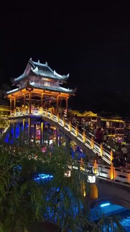 This video shows a nighttime scene of a traditional Chinese bridge adorned with lights, spanning over a body of water. People are walking across the bridge and gathered along the riverbank, suggesting a festive atmosphere. The architecture of the buildings in the background is also traditional, illuminated with warm lights, reflecting in the water. The sky is dark, with a full moon visible and some distant lights that could be lanterns or fireworks. The foreground includes some leafy branches, also illuminated with blue lights.