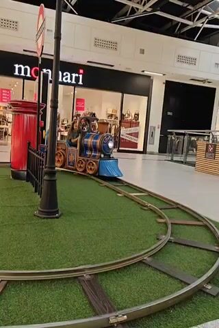 A toy train moves on a track in a mall. The train is blue and gold and has a light on the front. A child is sitting in the train. The train is on a fake grass track, and there is a lamppost and a red mailbox nearby. In the background, there are shops.