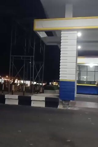 This video appears to be taken at night, likely in Tambontunong, Indonesia, given the context. The scene is a well-lit area, possibly a gas station or a similar commercial establishment, judging by the architectural elements and the presence of a security guard.

The main subject is a security guard walking across the frame. He is wearing a light-colored uniform shirt and dark pants, with a belt and possibly a badge. He is moving from left to right in the foreground. The guard is walking past a prominent pillar that is decorated with white horizontal lines and blue rectangular sections at its base. The pillar is part of a larger structure with a white and yellow canopy overhead, illuminated by lights.

In the background, other structures are visible, including a building with windows and what looks like an office or service area. There are also some vehicles and scaffolding in the distance, suggesting ongoing construction or maintenance. The ground in front of the structures is paved, with black and white striped barriers along the edge.

Further into the scene, a portable sign with text is visible. The text on the sign is in blue letters on a white background and reads "DILARANG MASUK TANPA IZIN," which translates to "NO ENTRY WITHOUT PERMISSION." This indicates a restricted area. There are also traffic cones in the background.

The overall atmosphere is calm and orderly, typical of a security presence at a commercial establishment during nighttime operations. The lighting suggests it is well after sunset, and the sky is dark. No specific weather conditions are apparent.