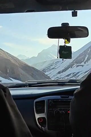The video captures a view from inside a moving car, looking out the front windshield at a mountainous landscape. The mountains are partially covered in snow. The road ahead is paved and has a guardrail visible in the foreground. The car's dashboard and rearview mirror are also visible. There is a faint green air freshener hanging from the rearview mirror. The sky is lightly colored, indicating possible early morning or late afternoon lighting.