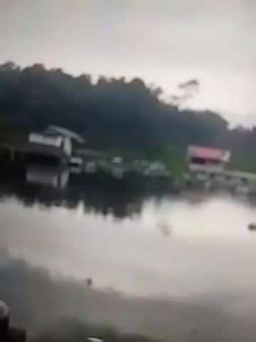 The video shows a body of water, likely a lake, with a wooded hillside in the background. There are a few structures on stilts over the water, one with a white roof and another with a red roof. The water is calm and reflects the overcast sky and the structures. There is some greenery visible along the shoreline. The camera appears to be positioned on a dock or boat, with a blurry railing visible in the foreground. The overall impression is a serene, natural landscape with some human habitation.