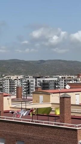A time-lapse video shows a rooftop view of a city with buildings, mountains in the background, and a cloudy sky. Laundry is hanging on clotheslines on several rooftops. The video progresses from left to right, panning across the scene.