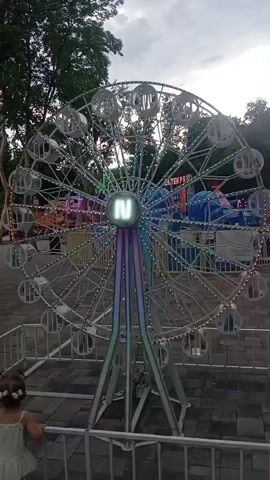 The video shows a Ferris wheel in what appears to be an amusement park or fairground. The Ferris wheel is illuminated with lights and has several cabins. In the foreground, a young child with light-colored hair tied in bows is standing behind a white fence, looking towards the Ferris wheel. Another person, mostly obscured by shadow and wearing a dark hooded garment and a black cap, is standing slightly behind and to the right of the child, also facing the Ferris wheel. The background features trees and other amusement park rides, suggesting an outdoor setting. The sky is overcast.