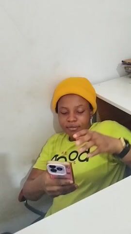 A young woman wearing a yellow beanie and a lime green t-shirt sits in front of a white desk and looks at her phone. She is holding the phone with both hands and her gaze is directed downwards. To her right, a stack of papers and books is piled on the desk. The background is a plain white wall.
