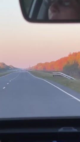 The video shows a view from the inside of a car looking forward at an empty two-lane road. The sky is a pale pink and orange, suggesting either sunrise or sunset. The sun is not directly visible. On the right side of the road, there is a line of trees with autumn foliage in shades of orange and red. On the left side of the road, there are also trees, but they appear greener and less vibrant. The road is paved and has white dashed lines marking the lanes. There are some distant lights visible on the horizon. In the rearview mirror at the top of the frame, a person wearing glasses is visible, presumably the driver. The overall impression is a peaceful drive through the countryside during a colorful time of day.