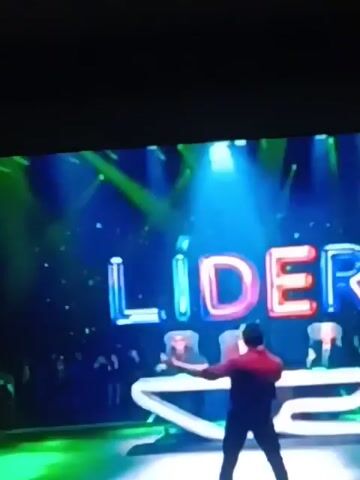 The video shows a man with a red shirt and black pants walking towards a stage with the word “Lider” spelled out in large, colorful letters behind him. The stage is lit with bright, colorful spotlights. The camera then cuts to a woman with curly blonde hair clapping, followed by a man with a beard wearing a red velvet blazer who is also clapping. The audience appears to be enjoying the performance or presentation on stage.