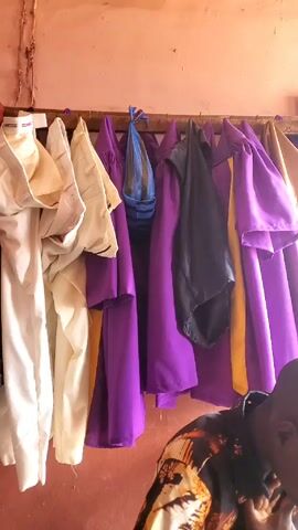 The video shows the interior of a small tailoring shop. The walls are painted red and there is a high shelf filled with piles of fabric in various colors and patterns. Below the shelf, a wooden table houses two vintage sewing machines. There is more fabric draped over the table and hanging near the sewing machines. In the initial shot, a rack of liturgical robes in colors like white, purple, and black hangs on the wall. The overall impression is that of a cluttered but functional workspace.