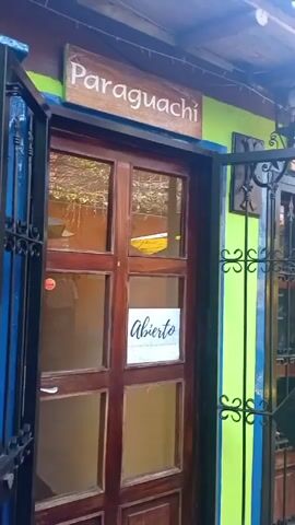 The video opens with a close-up of a vibrant storefront, showcasing a wooden door with glass panels and a sign above that reads "Paraguachí" in white letters on a light blue background. A smaller white sign on the door says "Abierto," meaning "Open." The wall of this shop is painted a bright lime green with a vertical blue stripe running down the side, next to a black wrought iron gate with decorative scrollwork.

As the camera pans to the right, it reveals another similar storefront next to the first one, also with a wrought iron gate and a sign above reading "La Mira." The walls maintain the same lime green and blue striped pattern. The camera continues to pan, showing a paved courtyard with terracotta-colored tiles underfoot. Overhead, a canopy of bare branches creates a dappled sunlight effect.

The next shop visible has a bright yellow wall with a blue stripe along the bottom. Another wooden door with glass panels is visible, and a sign above it appears to say "Guarame" and "Guacuco" (partially obscured). In front of this yellow shop, there's an outdoor seating area with a small round table and two yellow chairs, shaded by a large blue and yellow striped umbrella.

The pan continues, revealing an orange-painted wall. Beneath the orange wall, there's a blue-painted planter or fountain with a colorful, possibly tiled, base. Above it, a wooden sign reads "Fuente de los Deseos" (Wishing Fountain). To the right of the fountain, an open archway or entrance reveals more retail space with various colored T-shirts and clothing items displayed on racks and shelves. Another blue and yellow umbrella shades a second outdoor table, this one featuring a mosaic-like top with white floral patterns on a blue background. The video ends showing a closer view of the merchandise and the colorful umbrella.

The overall impression is a vibrant, colorful marketplace or small shopping area, likely in a warm climate, with an eclectic mix of shops and an inviting outdoor atmosphere.